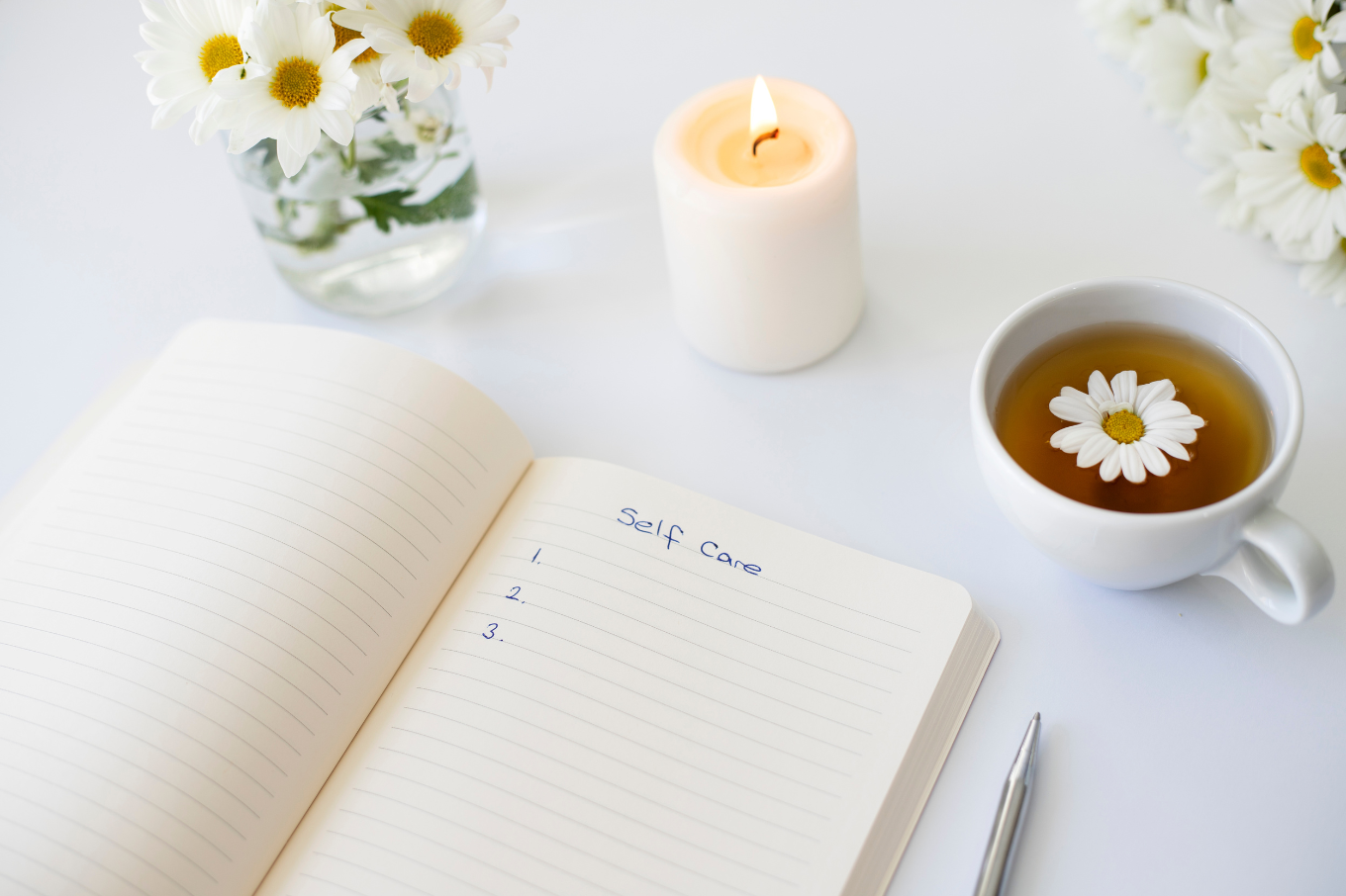 Close up of handwritten text "Self Care" in foreground with notebook, pen, cup of tea, flowers
